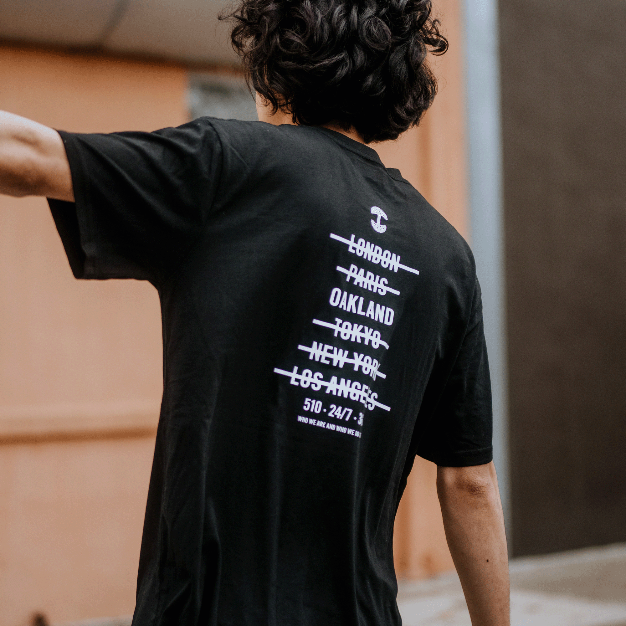 Seen from behind, a person with wavy hair wears Oaklandish's "Who We Are and Who We Do it For Tee," a classic fit black T-shirt made of 100% cotton. It features white crossed-out city names—London, Paris, Oakland, Tokyo, New York, Los Angeles—and "510-24/7-2" against peach urban walls.