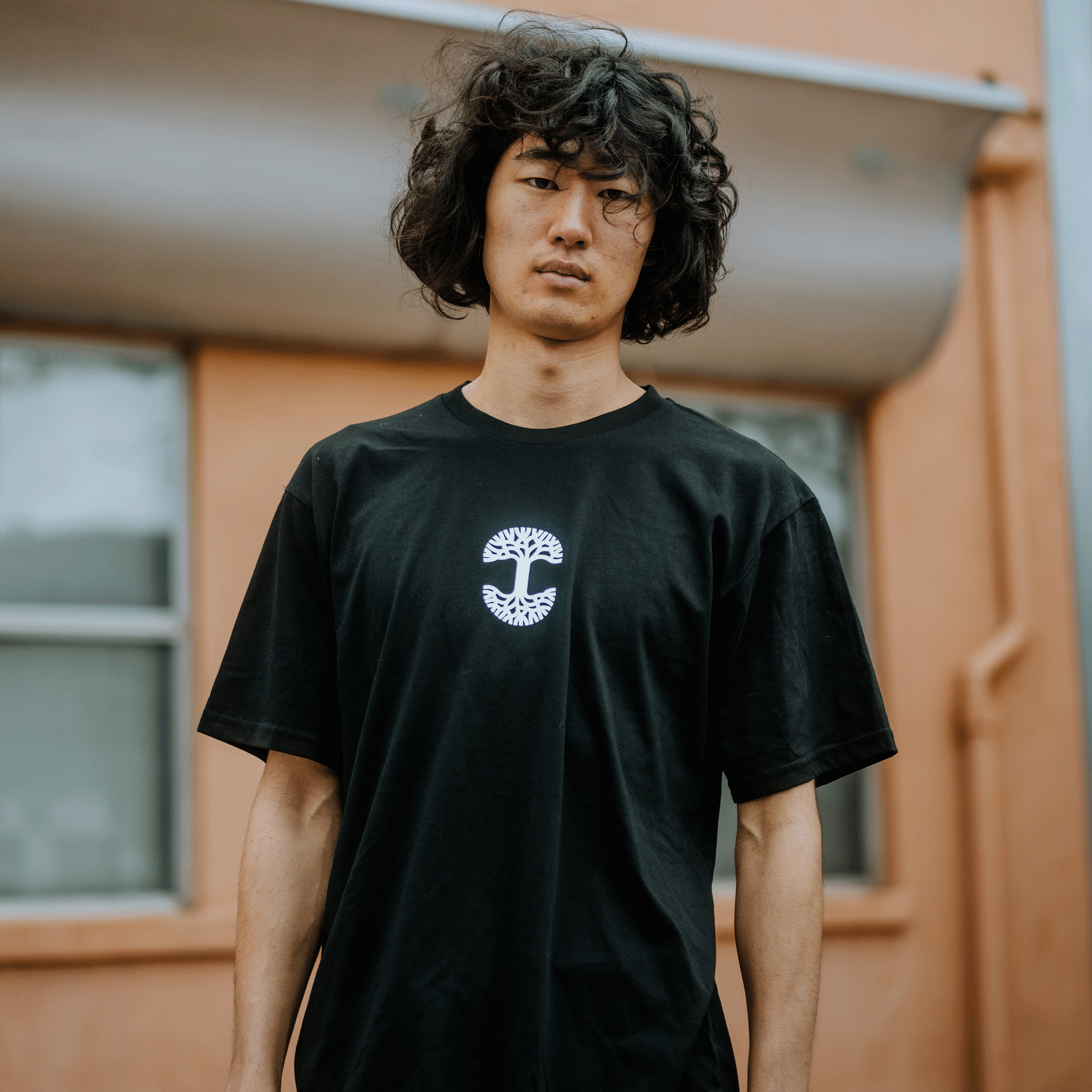 A person with curly hair stands outside wearing the "Who We Are and Who We Do it For Tee" by Oaklandish, featuring a white tree design on the front. The blurred orange wall and window suggest an Oakland vibe, while their neutral expression complements the casual urban setting.