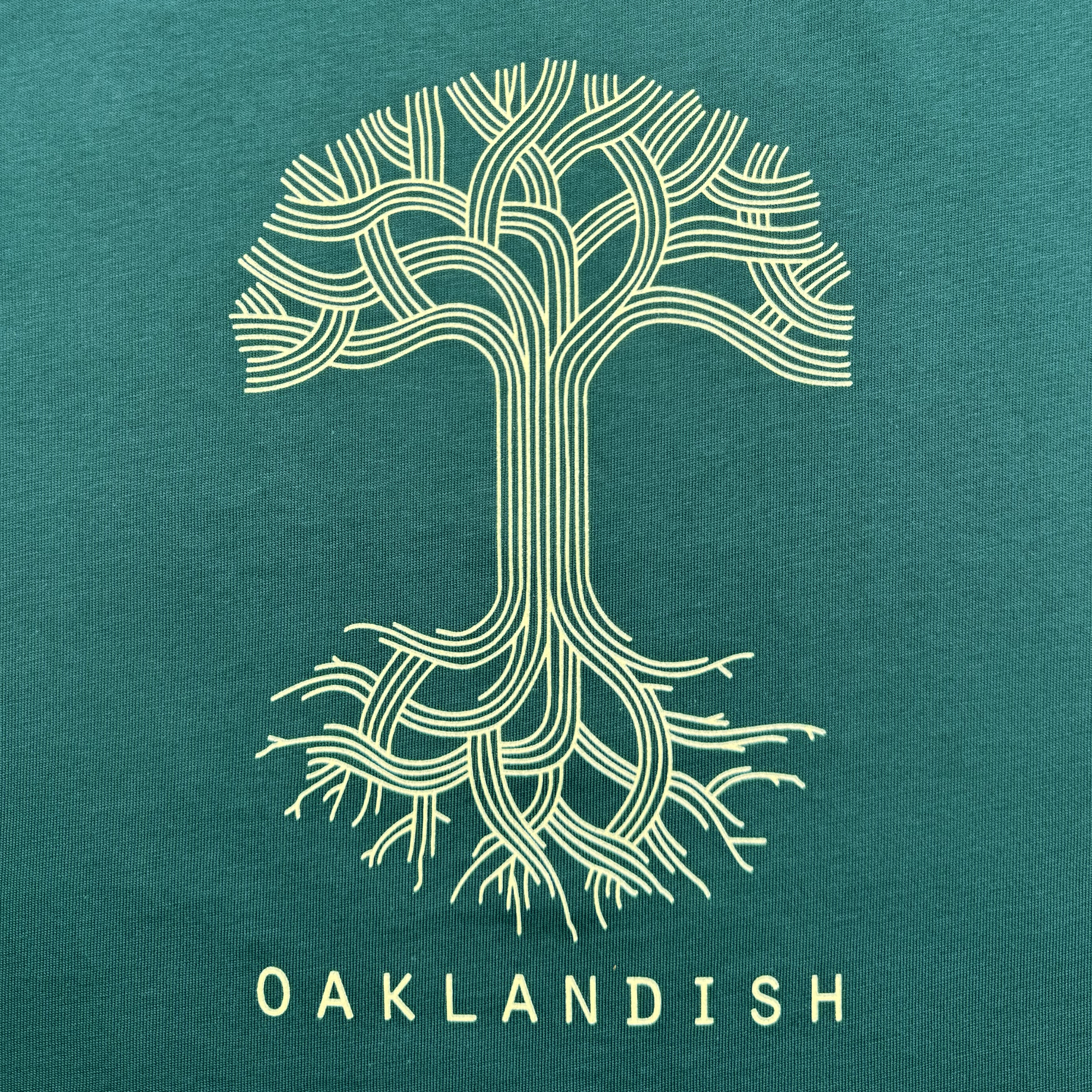 The Women's Oaklandish Classic Logo Tee features a stylized tree with intertwined branches and roots in line art on a dark green background, with "OAKLANDISH" below, all on a classic fit 100% cotton t-shirt.