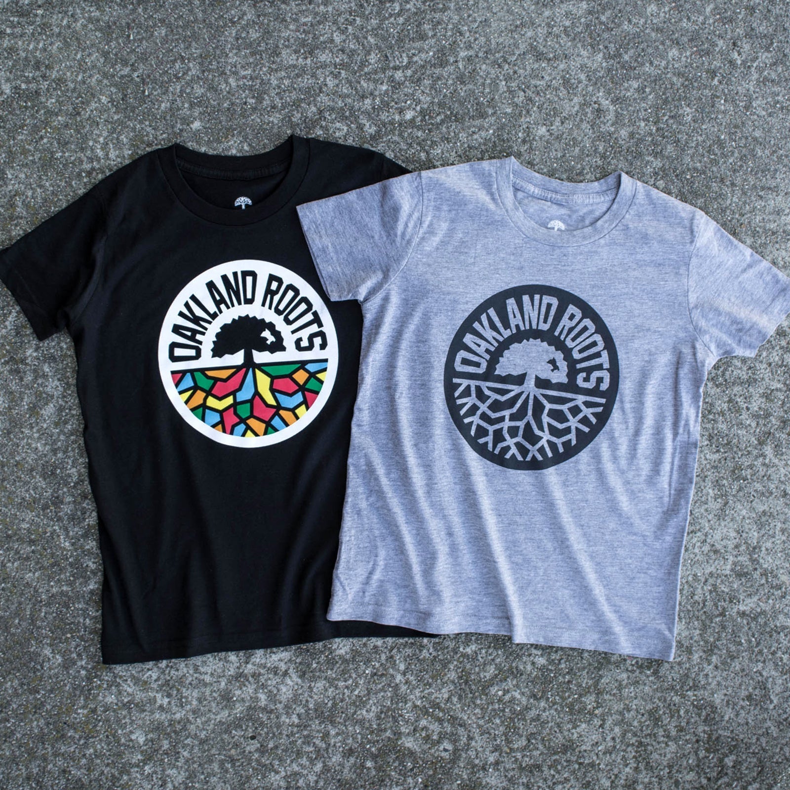 Two Youth Oakland Roots SC Classic Tees are displayed on a concrete surface. The black tee features a colorful tree logo with "OAKLAND ROOTS" in white text, inspired by the Oakland Roots Sports Club. The gray tee also showcases a tree logo with "OAKLAND ROOTS," but the design is monochromatic and exudes an Oaklandish vibe. Both logos depict a tree with visible roots underneath.