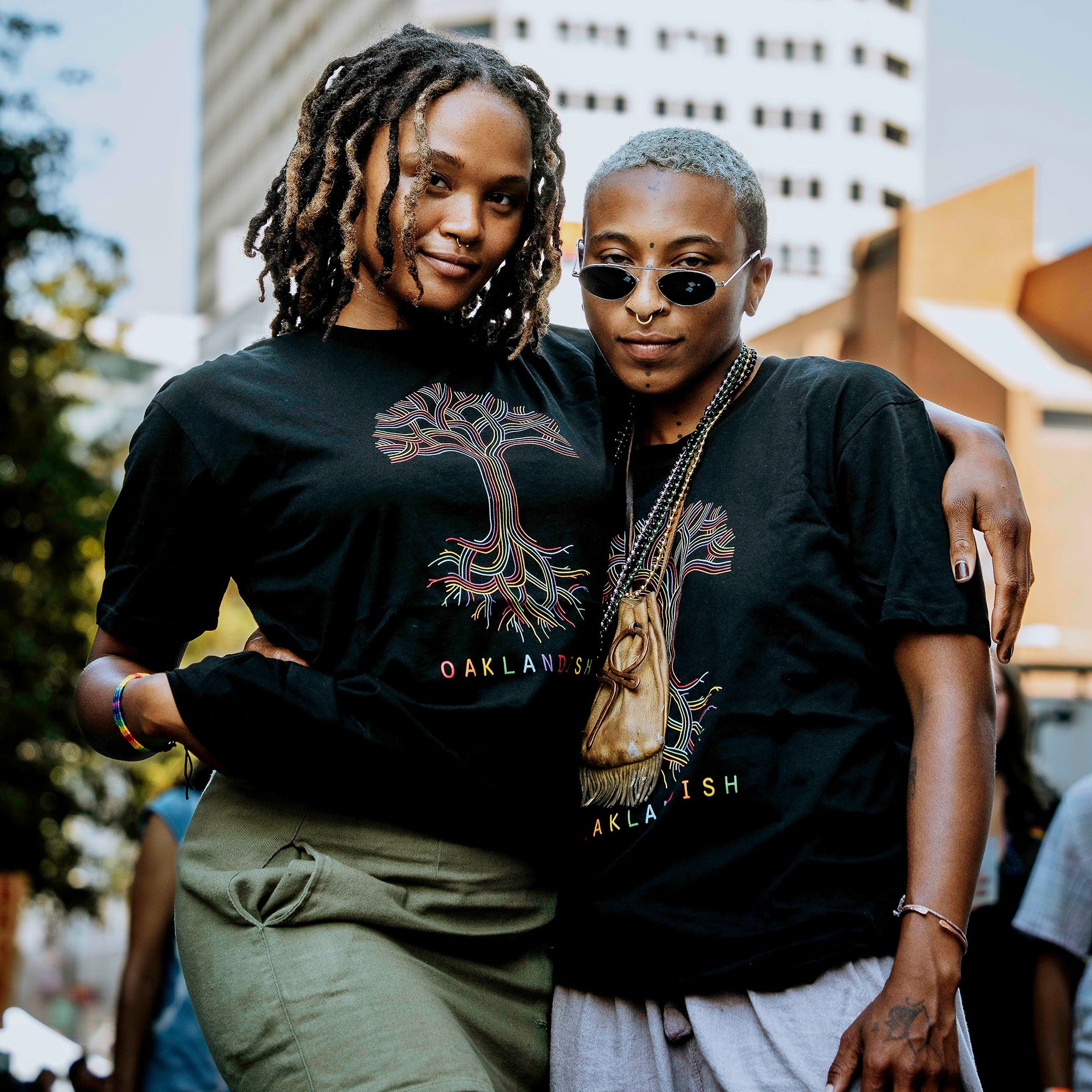 Two individuals stand closely together outdoors, both wearing the Oaklandish Pride Classic Logo Tee, featuring a limited-edition black design with a colorful tree graphic and the word "OAKLANDISH" printed below, proudly showcasing their Oakland spirit. One has short blonde hair and wears round sunglasses, while the other has long braided hair. Urban buildings form the background.